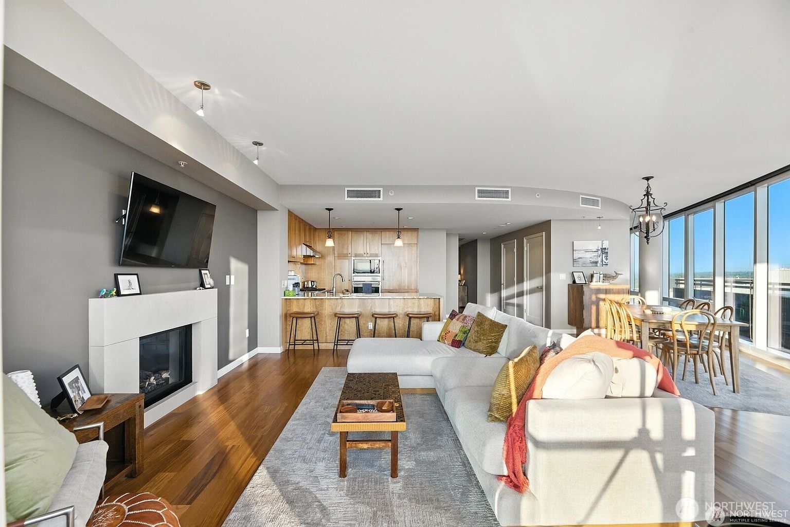This is an interior shot of a modern living room, showcasing an open-concept design that integrates the living area, kitchen, and dining space. The living room features a large sectional sofa, a fireplace with a mounted TV above, and a stylish coffee table on a light-colored rug. The kitchen has wooden cabinetry and bar seating, while the dining area is illuminated by a chandelier and natural light from large windows, creating a bright and inviting atmosphere.