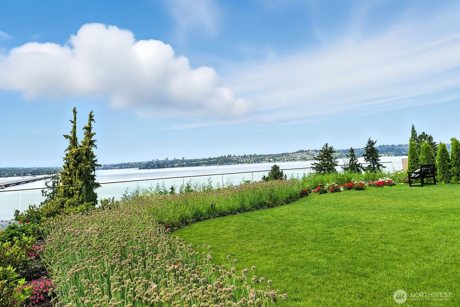 This image showcases a beautifully landscaped yard with a lush green lawn, a variety of flowering plants, and mature trees. A bench is situated on the lawn, offering a place to relax and enjoy the view of the water and distant cityscape. The overall impression is one of tranquility and natural beauty, highlighting the property's outdoor living space.