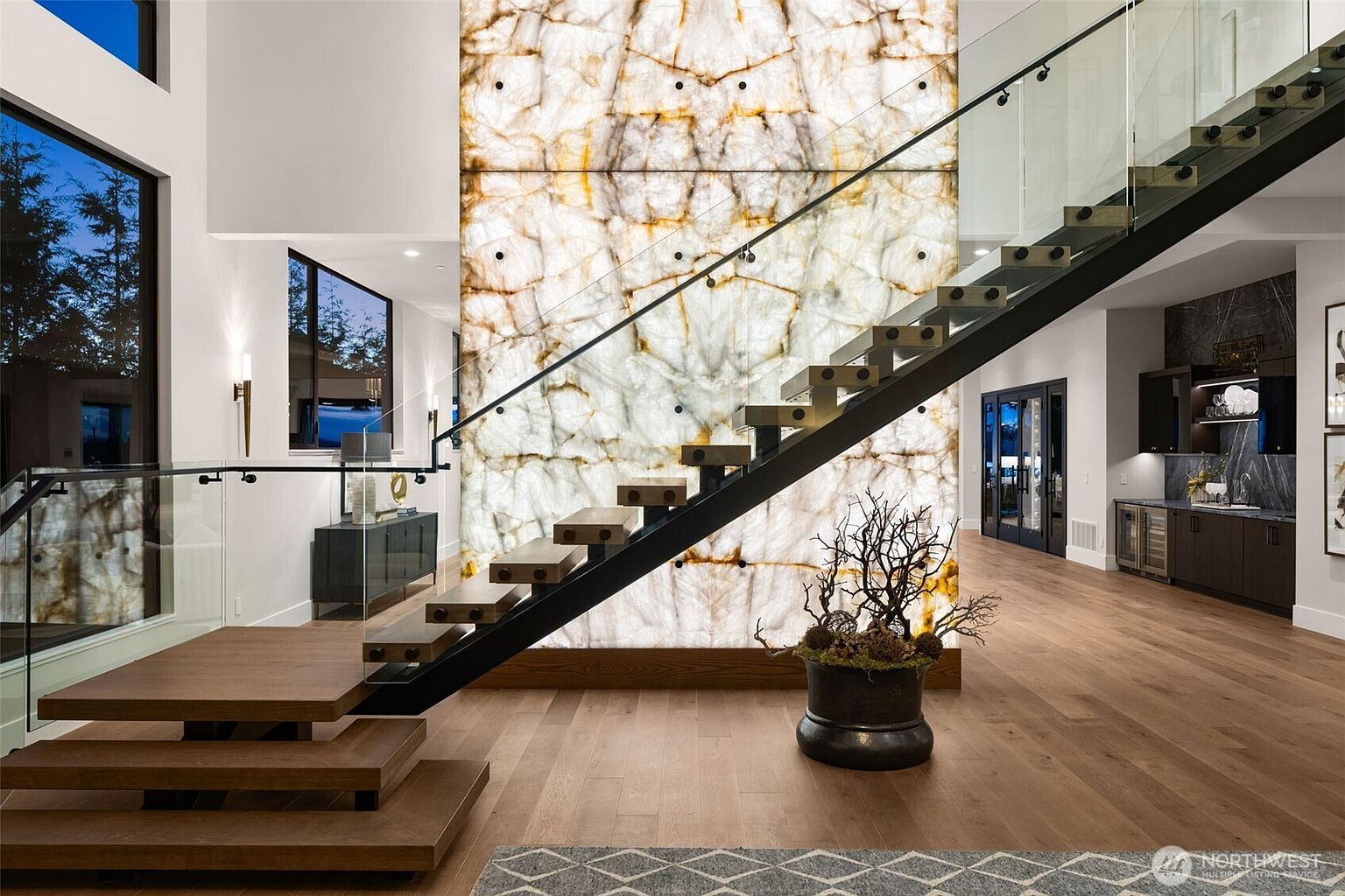 This interior shot showcases a modern hallway and staircase with a unique design. The floating wooden stairs are accented by a black metal frame and glass railings, while a striking backlit stone wall adds a luxurious touch. The open layout and hardwood flooring contribute to a spacious and elegant atmosphere.