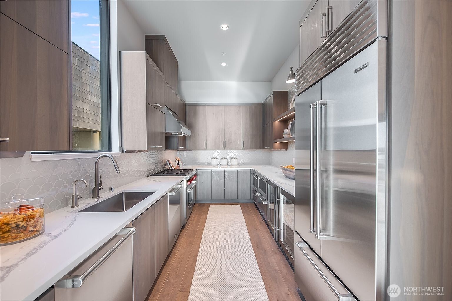 This is a modern kitchen with sleek, light wood cabinetry and stainless steel appliances, including a large refrigerator. The countertops are white with subtle veining, and the backsplash features a decorative tile pattern. A long runner rug adds warmth to the hardwood floors, and recessed lighting illuminates the space, creating a clean and sophisticated atmosphere.