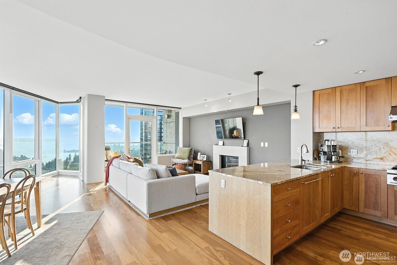 This is an interior shot of a modern living room and kitchen area. The living room features a large sectional sofa, floor-to-ceiling windows with a view, and a fireplace. The kitchen has wooden cabinets, granite countertops, and an island with a sink. The overall impression is bright, open, and luxurious.