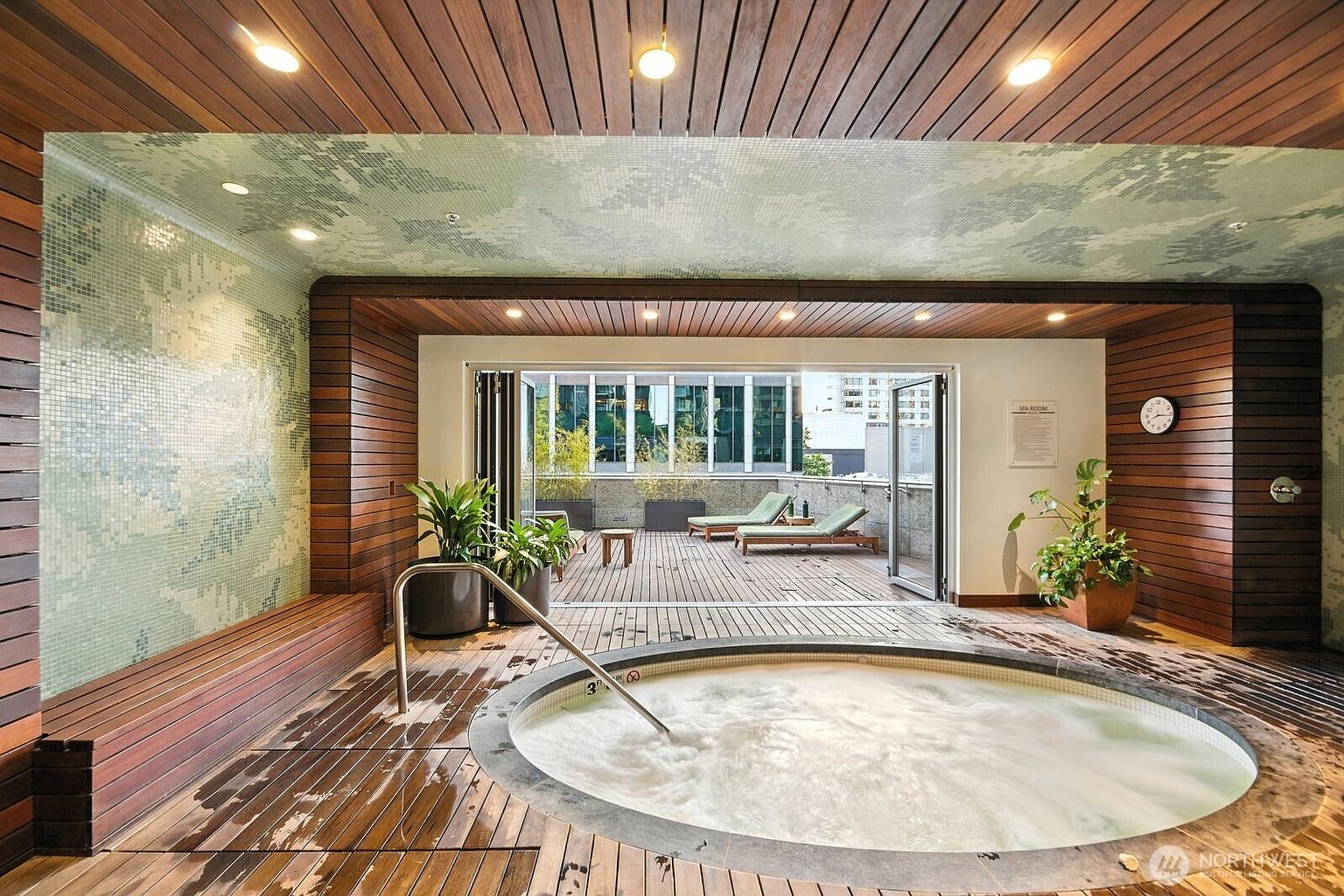 This image showcases an indoor spa area featuring a circular hot tub with bubbling water, surrounded by a wooden deck. Large folding doors open to an outdoor patio with lounge chairs, creating a seamless indoor-outdoor experience. The space is decorated with plants and features wood paneling and mosaic tile accents, giving it a luxurious and relaxing ambiance.