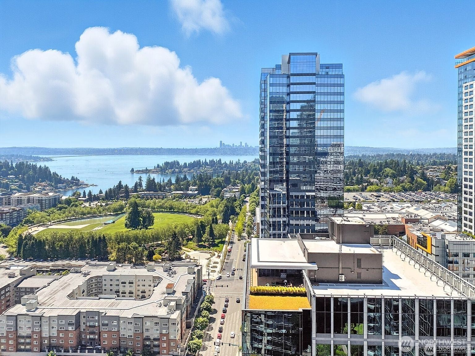 This aerial view showcases a modern cityscape with a prominent glass skyscraper reflecting the sky. Lush green spaces and a large lake provide a scenic backdrop, while well-maintained streets and surrounding buildings suggest a vibrant urban environment. The image conveys a sense of luxury and prime real estate location.