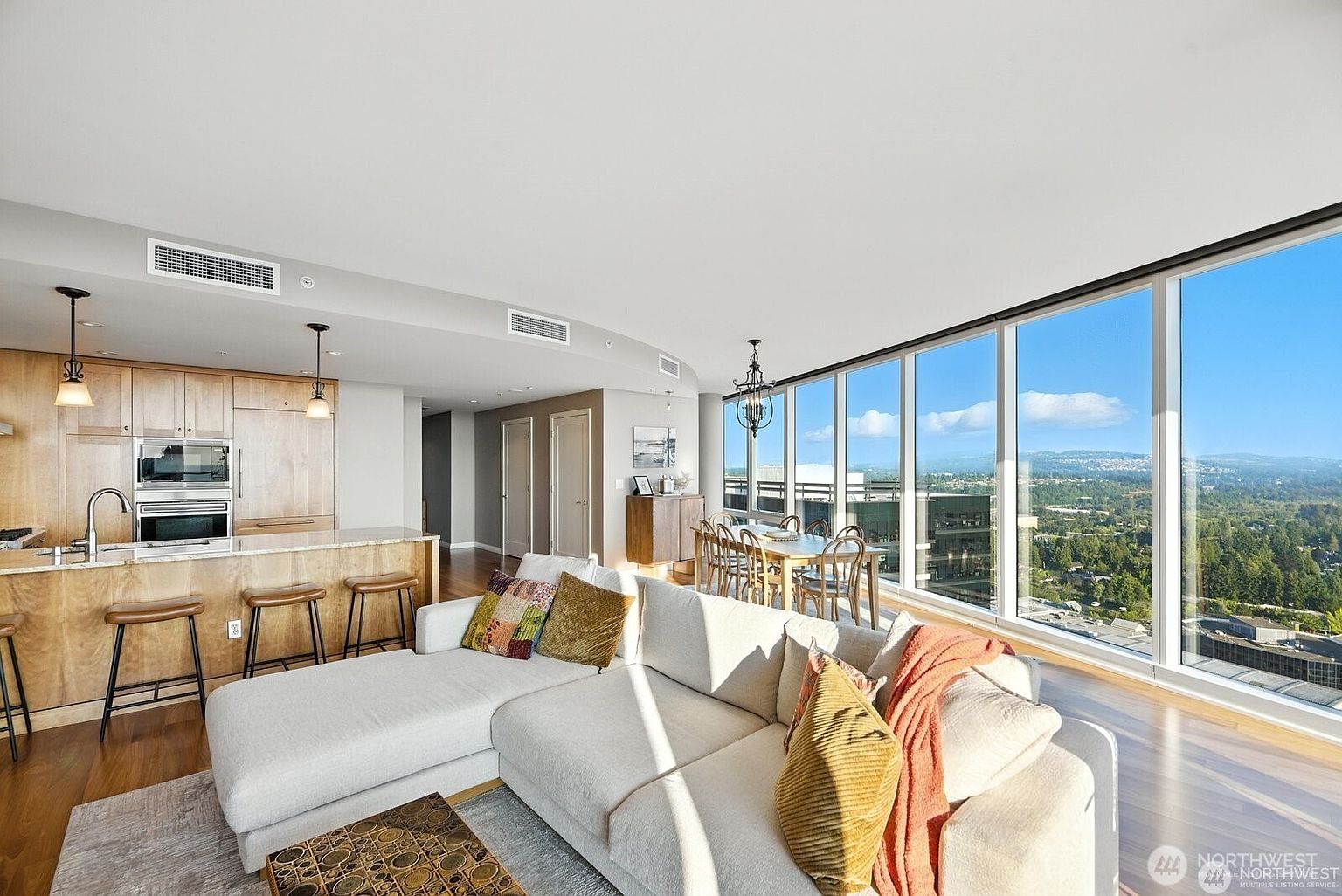 This is a bright and airy living room in a high-rise apartment, featuring a large sectional sofa with decorative pillows and a unique coffee table. The open-concept design seamlessly integrates the kitchen and dining area, while floor-to-ceiling windows offer panoramic views of the city and surrounding landscape. The hardwood floors and modern chandelier add a touch of elegance to the space.