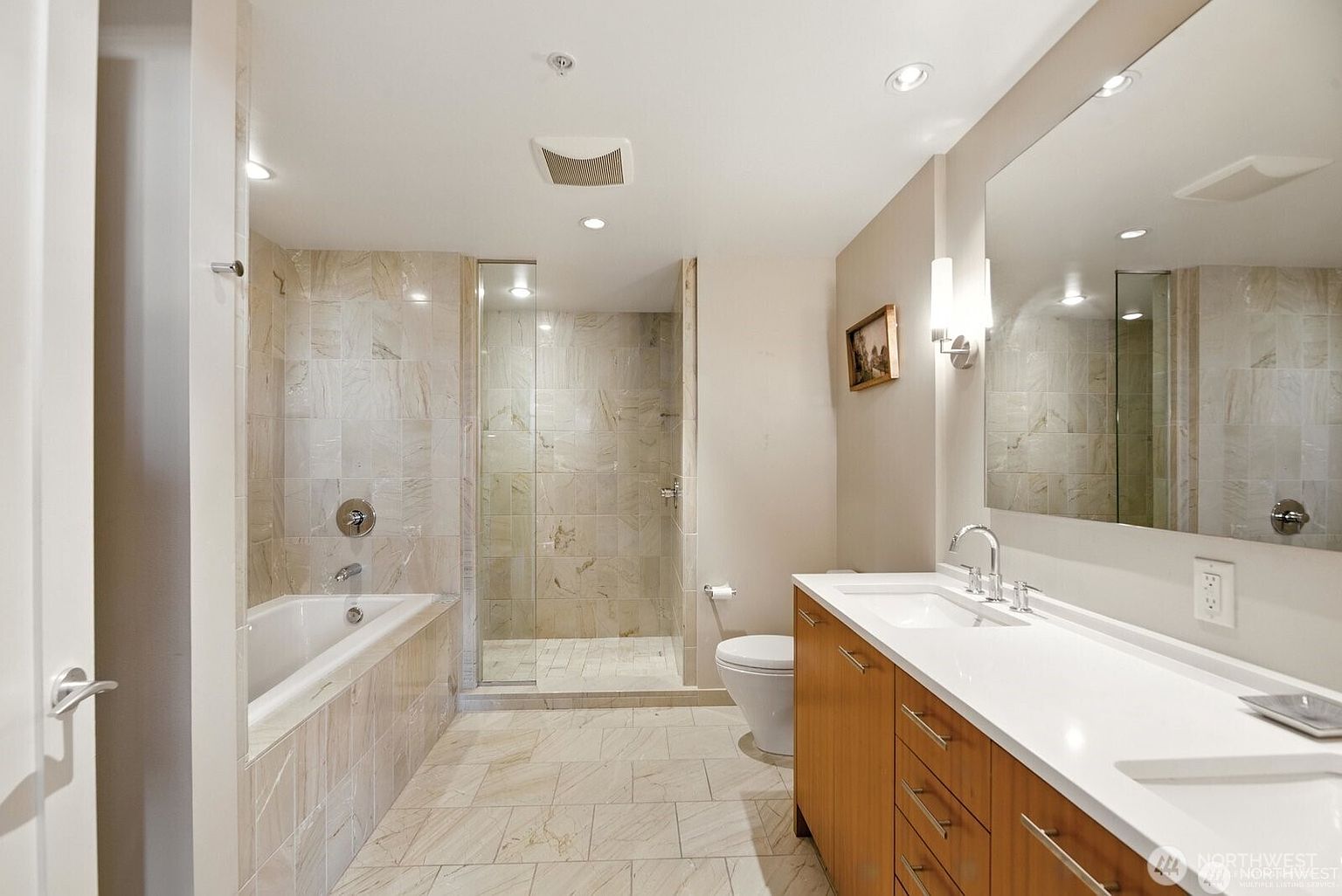 This is a well-lit primary bathroom featuring a bathtub and a separate glass-enclosed shower. The vanity has a white countertop and wood-finish cabinets, complemented by a large mirror. The bathroom is tiled with light-colored stone, creating a clean and luxurious feel.