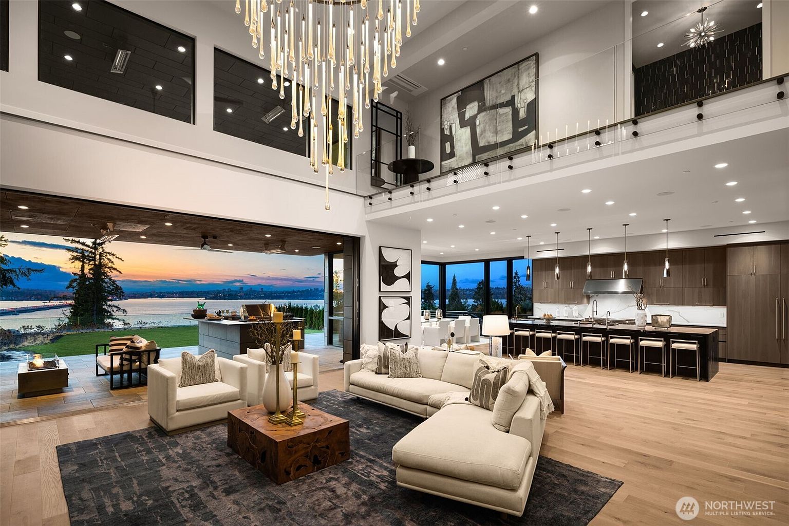 This is a spacious, modern living room with an open floor plan that seamlessly connects to the kitchen and outdoor patio. The room features high ceilings, a large chandelier, and expansive windows offering stunning views of the water. The neutral color palette, comfortable seating, and stylish decor create an inviting and luxurious atmosphere, perfect for entertaining or relaxing.