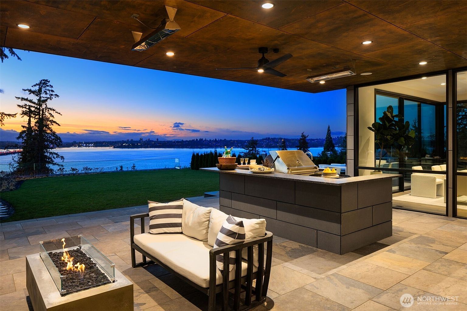 This image showcases a luxurious outdoor patio area with a stunning waterfront view at dusk. The space features a built-in outdoor kitchen with a grill, a modern fire pit, and comfortable seating. The patio is paved with stone tiles, and the ceiling is equipped with fans and heaters, creating a perfect setting for relaxation and entertainment.