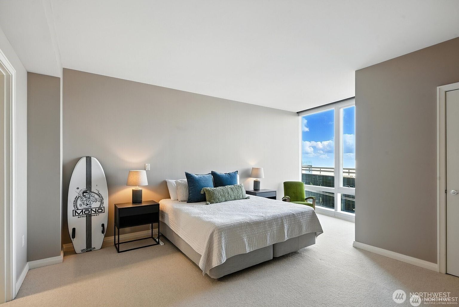 This is a bright and airy primary bedroom featuring a large window with a city view. The room is decorated in neutral tones with pops of blue and green in the pillows. A surfboard leans against the wall, adding a unique touch to the space. The overall impression is clean, modern, and inviting.