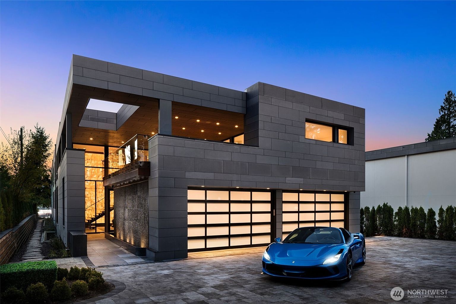 This is a striking front view of a modern, multi-level home featuring a sleek, dark gray exterior with contrasting light from the garage doors and interior spaces. A blue sports car is parked in the paved driveway, adding a touch of luxury. The architectural design emphasizes clean lines and geometric shapes, creating a sophisticated and contemporary aesthetic.