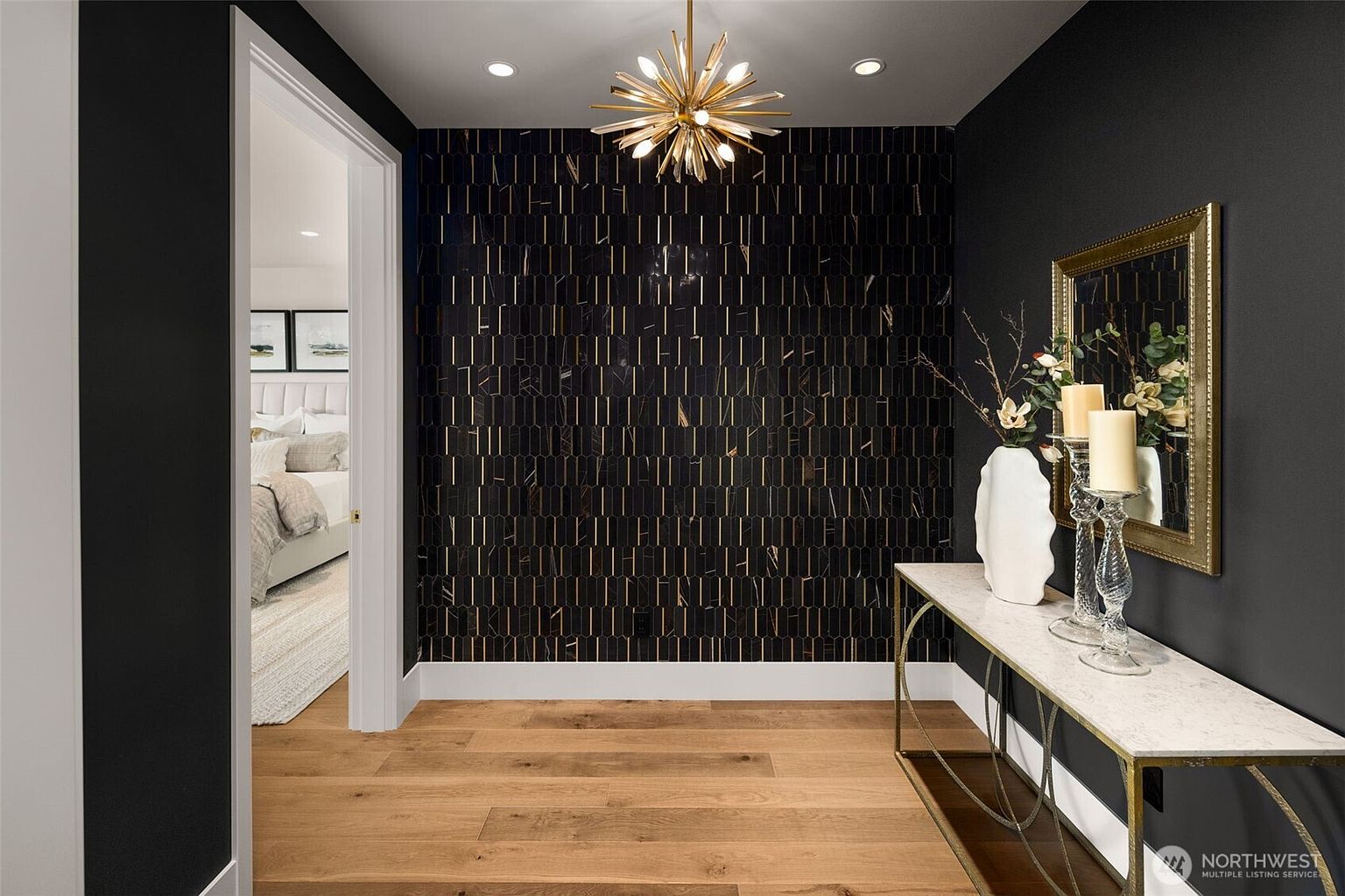 This interior shot showcases a stylish hallway with a striking black and gold accent wall, complemented by warm wood flooring. A modern chandelier adds a touch of elegance, while a console table with decorative items enhances the sophisticated ambiance. The open doorway provides a glimpse into an adjacent room, creating a sense of depth and flow.