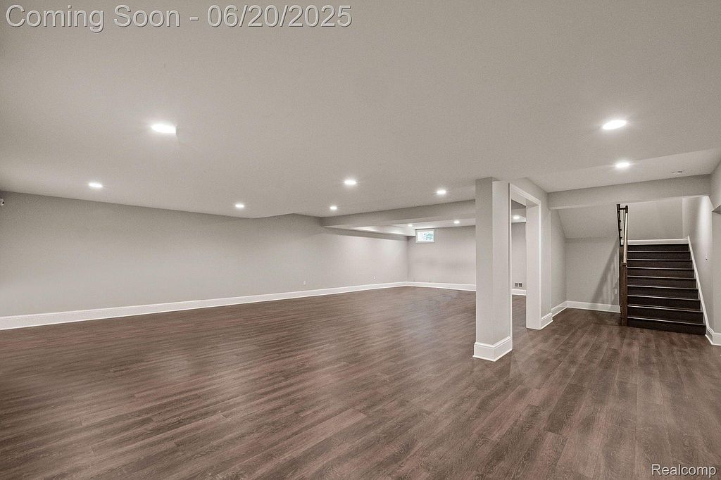 This is a spacious, newly finished basement with modern dark wood-look flooring and neutral gray walls. Recessed lighting provides ample illumination, and white trim adds a clean, contemporary touch. The area includes support columns and access to stairs, suggesting a versatile space for recreation or storage.