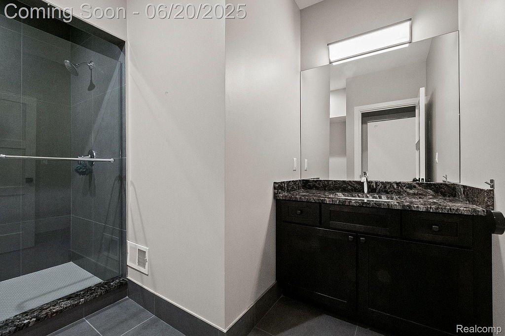This is a modern bathroom featuring a dark vanity with a granite countertop and a large mirror. A glass-enclosed shower with gray tiling is visible to the left. The room has a clean and contemporary aesthetic, suitable for a guest or secondary bathroom.