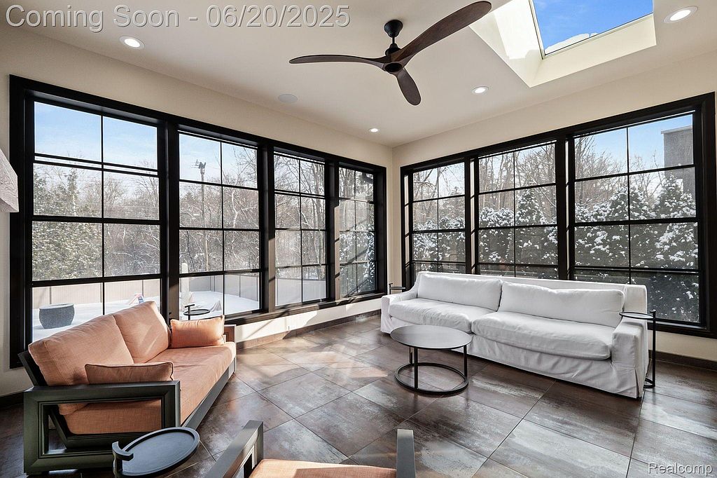 This sunroom features large windows with black frames, providing ample natural light and views of the snowy outdoors. The room is furnished with a light orange sofa and a white sofa, along with a round coffee table. The flooring is tiled, and a ceiling fan adds to the room's comfort.