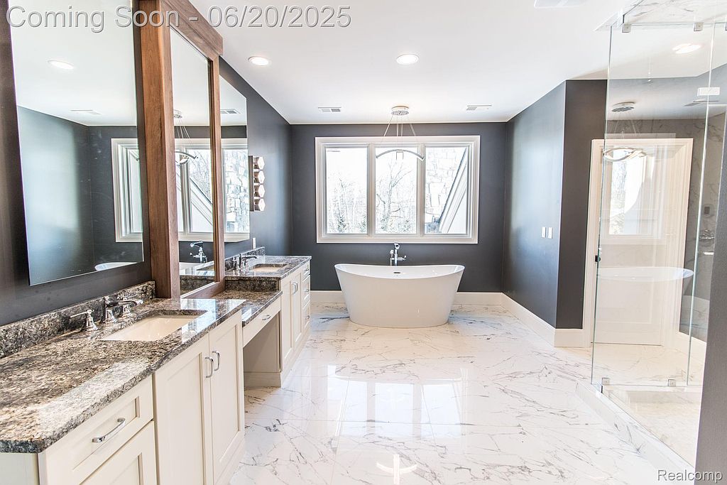 This is a spacious primary bathroom featuring a double vanity with granite countertops, a freestanding bathtub under a large window, and a glass-enclosed shower. The flooring is marble, and the walls are painted a dark gray, creating a luxurious and modern feel. The room is well-lit and appears clean and well-maintained.