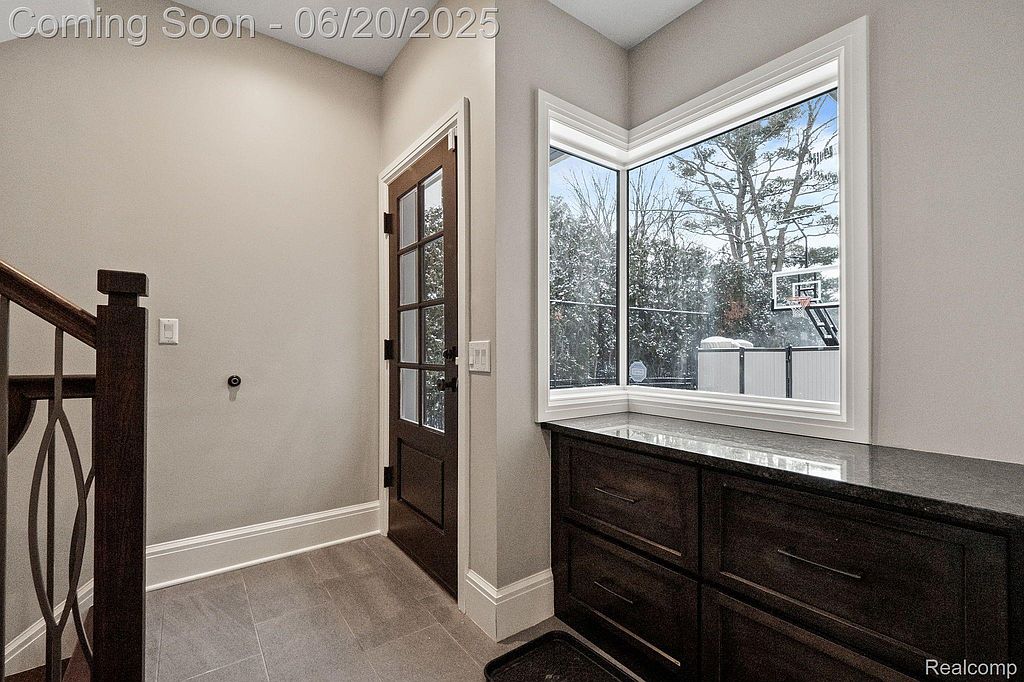 This interior shot showcases a hallway area with a dark wood door featuring glass panes, leading to the outside. A built-in dark wood cabinet with a granite countertop sits beneath a large window offering a view of the outdoors, including a basketball hoop. The flooring is a neutral tile, and the walls are painted in a light, warm tone, creating a welcoming and functional space.