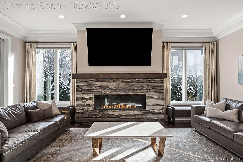 This living room features a modern fireplace with a stone surround and a dark wood mantel, flanked by windows with sheer curtains. Two leather sofas face each other across a light-colored marble coffee table, creating a symmetrical and inviting space. The room is well-lit and appears spacious, with a neutral color palette.