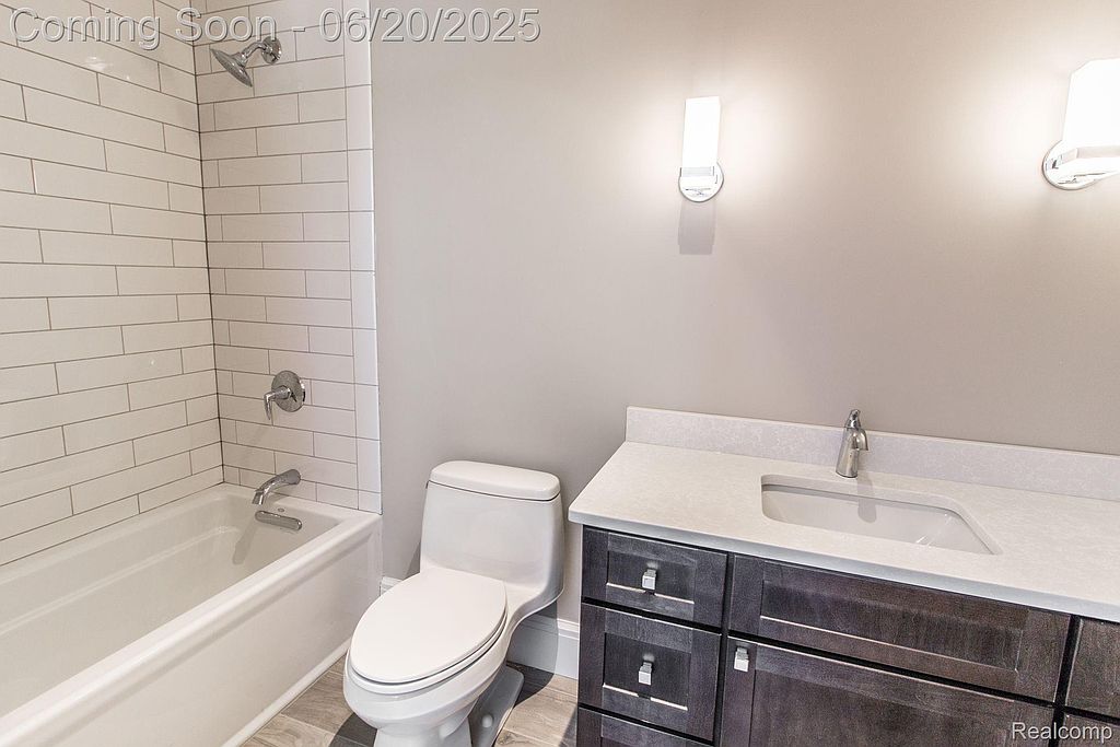 This is a clean and modern bathroom featuring a white subway tile shower/tub combination, a white toilet, and a dark wood vanity with a white countertop and sink. The walls are painted a neutral gray, and the lighting is provided by modern sconces. The overall impression is fresh and updated.