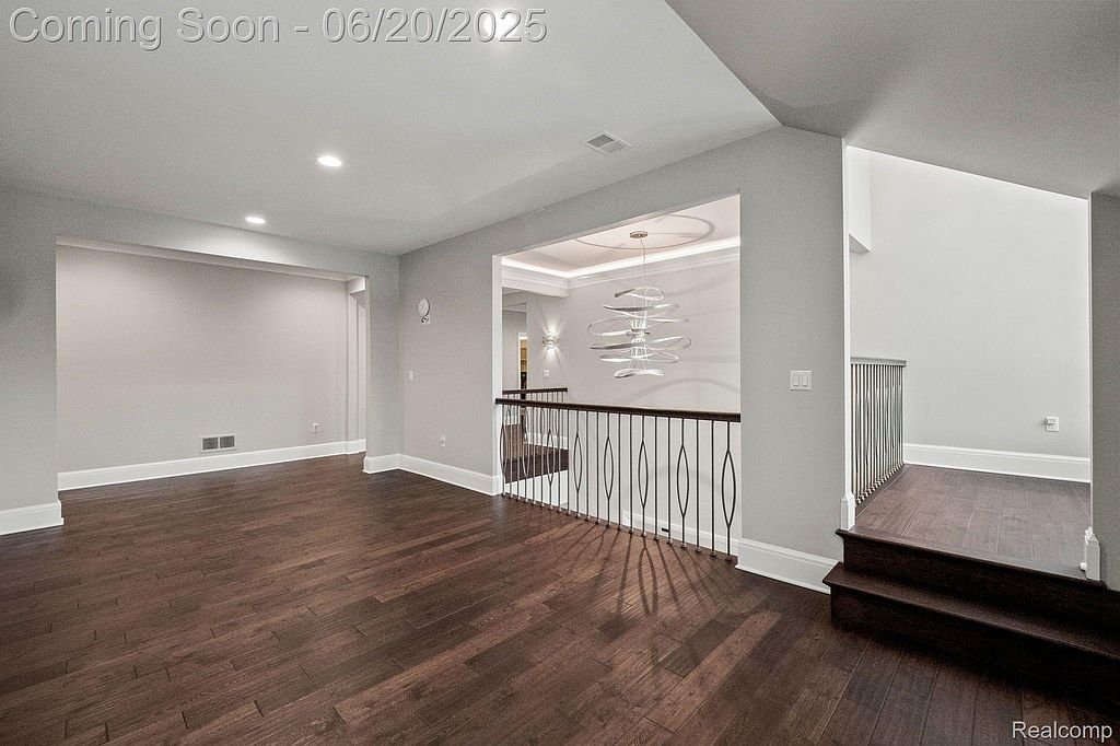 This interior shot showcases a modern living room with dark hardwood floors and light gray walls. The open layout leads to a hallway with decorative railing and a glimpse of a stylish chandelier. The space is well-lit and appears spacious, creating an inviting atmosphere.