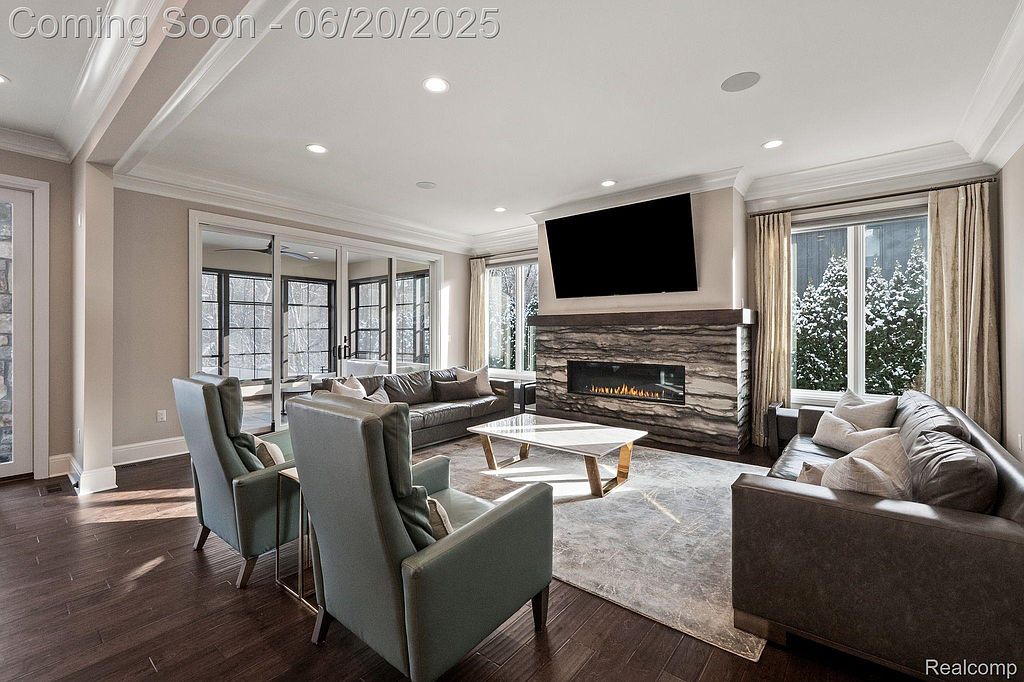 This is a well-lit living room featuring hardwood floors, a modern fireplace with a mounted television, and comfortable seating arrangements. The room has large windows that provide natural light and a view of the outdoors, enhancing the inviting and luxurious atmosphere. The neutral color palette and elegant furnishings contribute to a sophisticated and comfortable living space.