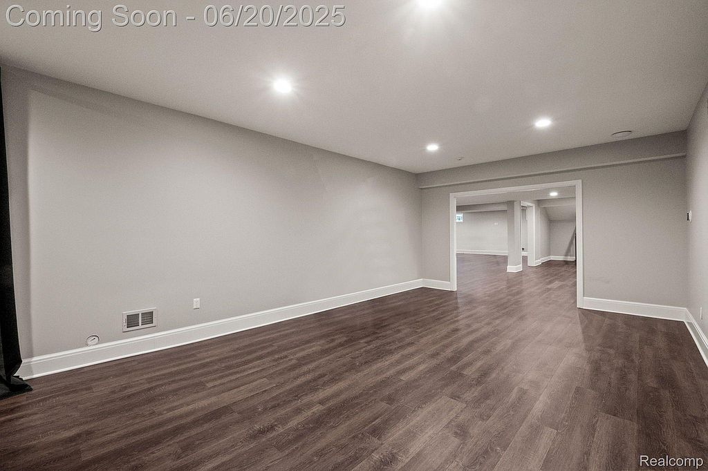 This is a spacious, newly finished basement with dark wood-look flooring and neutral gray walls. Recessed lighting provides ample illumination, and a large opening leads to another area, suggesting a versatile layout. The room is clean and modern, ready for various uses.