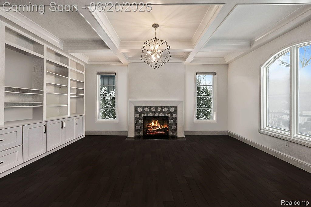 This is a well-lit living room featuring a fireplace, built-in shelving, and dark hardwood floors. The room has a modern yet cozy feel, with a geometric chandelier adding a touch of elegance. Large windows provide natural light and a view of a snowy landscape.