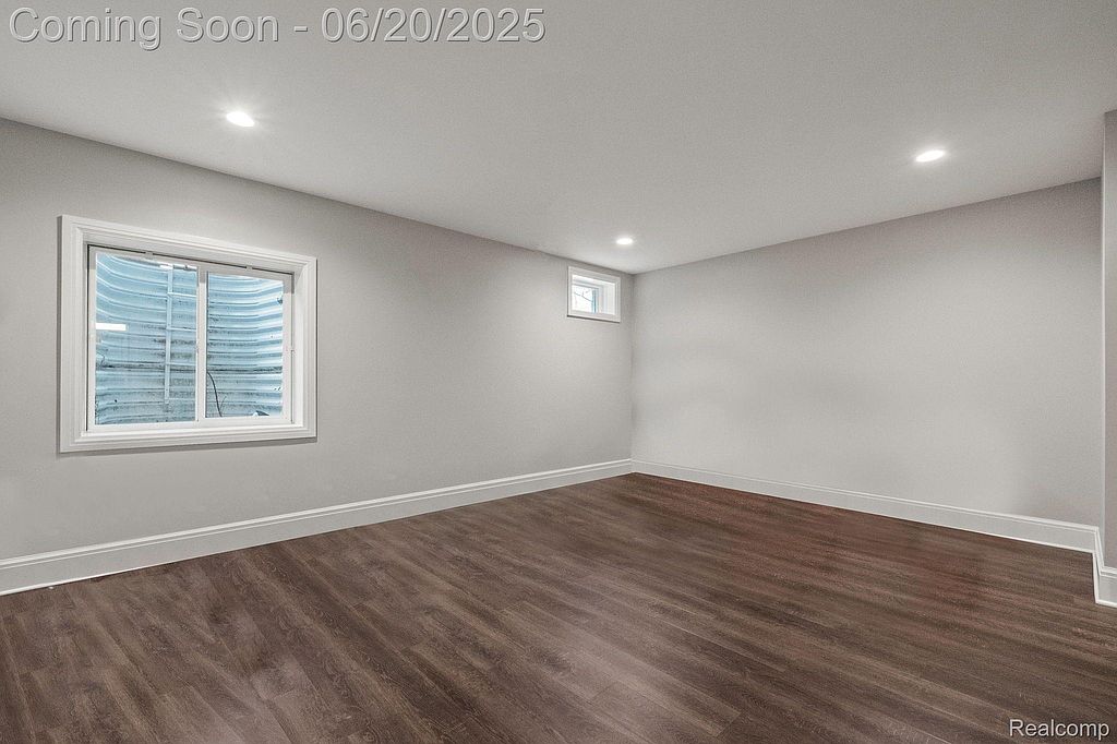 This is a clean, finished basement space with dark wood-look flooring and light gray walls. A window well provides natural light, and recessed lighting illuminates the room. The space appears versatile and ready for various uses, such as a recreation room or home office.