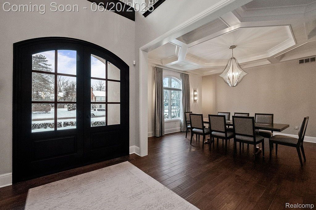 This image showcases a dining room with dark hardwood floors, a coffered ceiling with a modern chandelier, and a large dining table with upholstered chairs. A set of arched windows with curtains provides natural light, and a dark-framed glass door is visible to the left. The room exudes elegance and sophistication, making it ideal for formal gatherings.