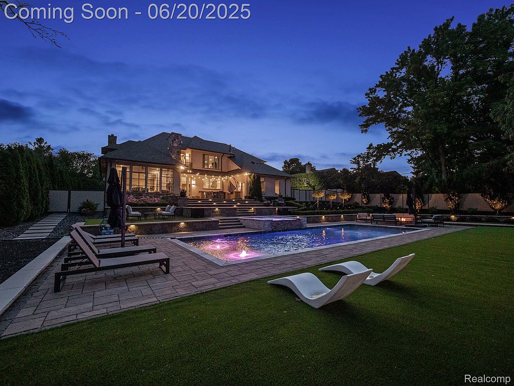 This image showcases a luxurious backyard featuring a stunning pool and spa area. The pool is surrounded by elegant patio stones and lush green grass, complemented by modern lounge chairs. The backdrop includes a beautifully lit house with outdoor seating, creating an inviting and upscale ambiance perfect for relaxation and entertainment.