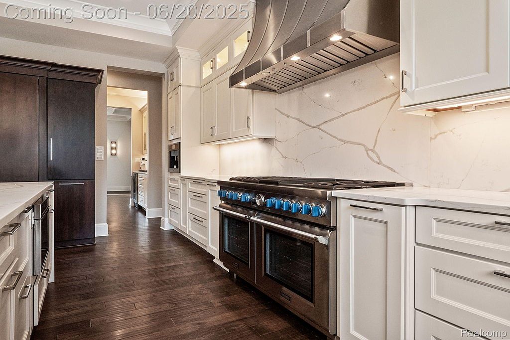 This is a well-lit kitchen featuring white cabinetry, stainless steel appliances, and dark hardwood floors. The kitchen includes a large range with a stainless steel hood and a built-in coffee maker. The overall impression is modern and luxurious.