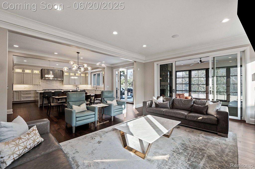 This is an open-concept living room and kitchen area featuring hardwood floors, neutral walls, and recessed lighting. The living room has a large gray sofa, two blue armchairs, and a modern coffee table, while the kitchen boasts stainless steel appliances and a spacious island with bar seating. Large sliding glass doors provide ample natural light and access to an outdoor space.