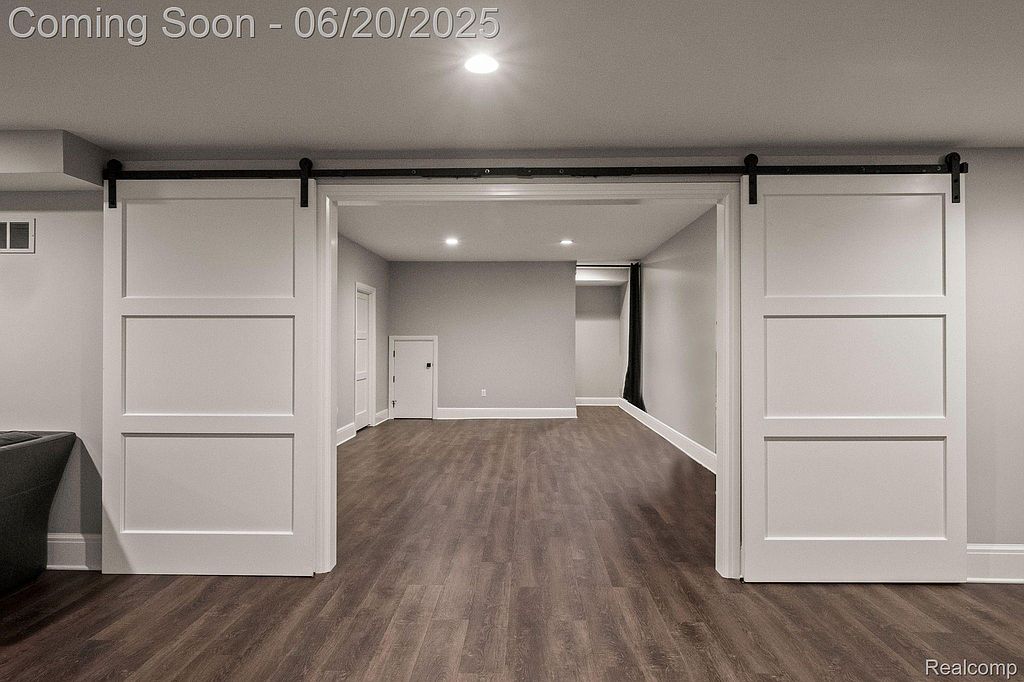 This image showcases a finished basement with modern sliding barn doors leading into the space. The flooring is a dark wood laminate, and the walls are painted in a neutral gray tone. The room is well-lit with recessed lighting, creating a clean and inviting atmosphere.