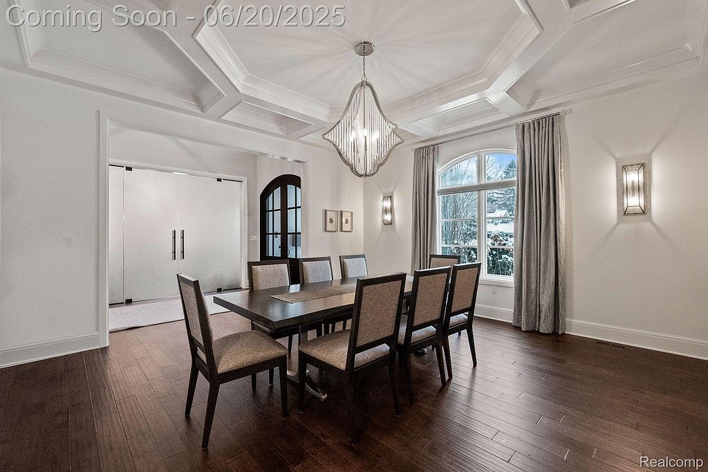 This is an elegant dining room featuring a dark wood table with seating for eight, illuminated by a modern chandelier. The room has a sophisticated feel with its coffered ceiling, hardwood floors, and large window with drapery. The space is well-lit and appears spacious, perfect for formal dining.