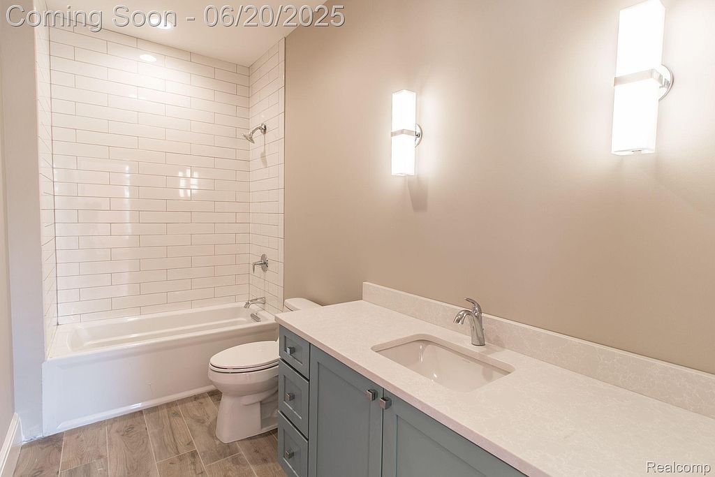 This is a well-lit bathroom featuring a bathtub with white subway tile surround, a toilet, and a vanity with a light blue cabinet and white countertop. The walls are painted a neutral beige, and the flooring appears to be wood-look tile. The overall impression is clean and modern.