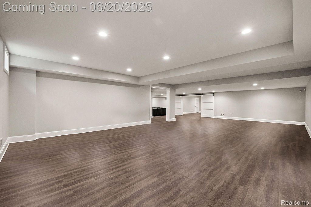 This is a spacious, finished basement with modern dark wood-look flooring and neutral gray walls. Recessed lighting provides ample illumination, and there are doorways leading to other rooms, including one with sliding barn doors. The space appears clean and well-maintained, offering potential for various uses.