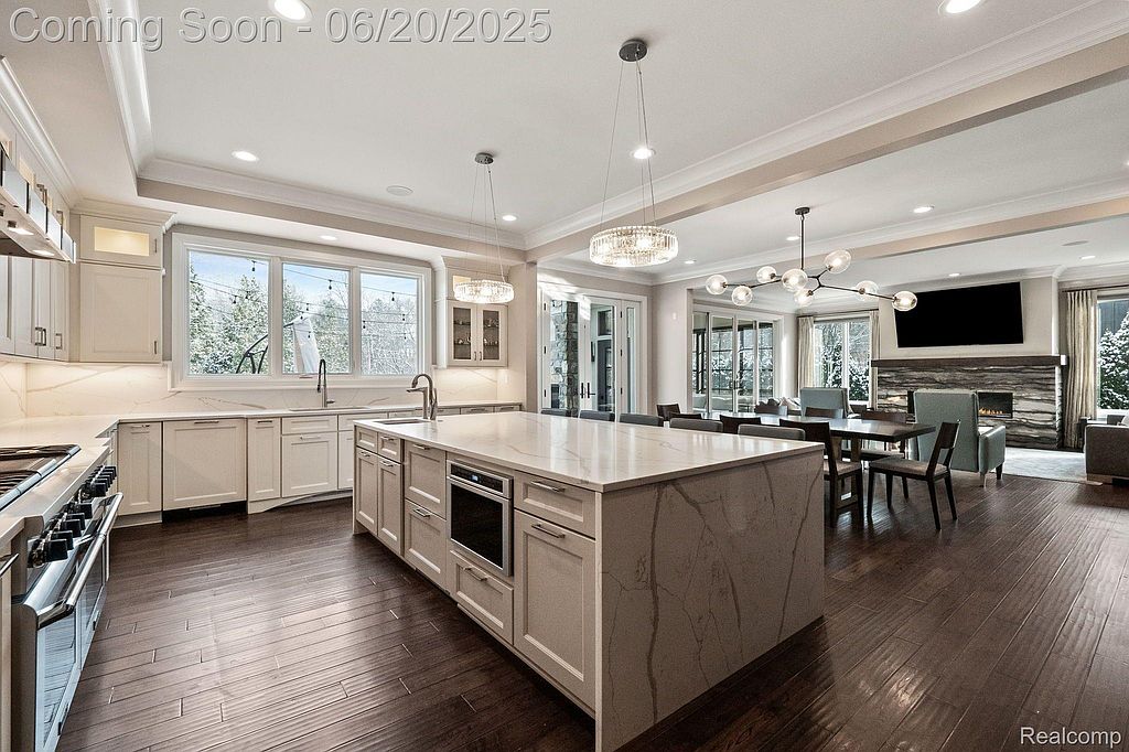 This interior shot showcases a modern, open-concept kitchen and dining area. The kitchen features white cabinetry, a large island with a light countertop, and stainless steel appliances. The dining area is adjacent, with a view of the fireplace and television, creating a seamless and inviting living space.