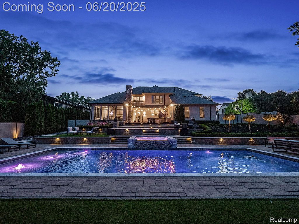 This image showcases a luxurious backyard featuring a large swimming pool and spa area. The pool is illuminated with vibrant lighting, and the surrounding patio is well-manicured with lush landscaping. The backdrop includes a grand house with outdoor seating areas, creating an inviting and upscale outdoor living space.