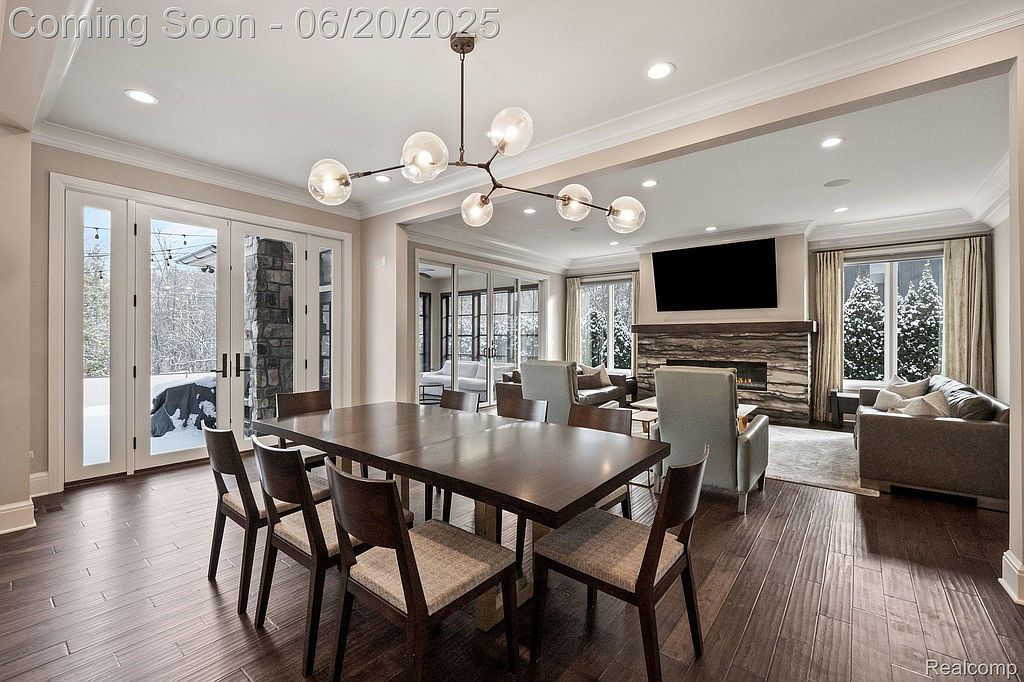 This is an interior shot of a dining room that seamlessly transitions into a living room. The dining area features a large wooden table with chairs and a modern chandelier, while the living room boasts a fireplace, a large television, and comfortable seating. The hardwood floors and neutral color palette create a warm and inviting atmosphere.
