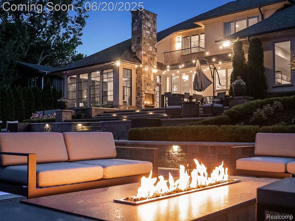 This image showcases a luxurious outdoor living space with a modern fire pit as the focal point. Comfortable seating surrounds the fire, and the patio extends to a multi-level deck with outdoor dining and lounge areas. String lights add a warm ambiance to the well-manicured backyard, creating an inviting atmosphere for relaxation and entertainment.