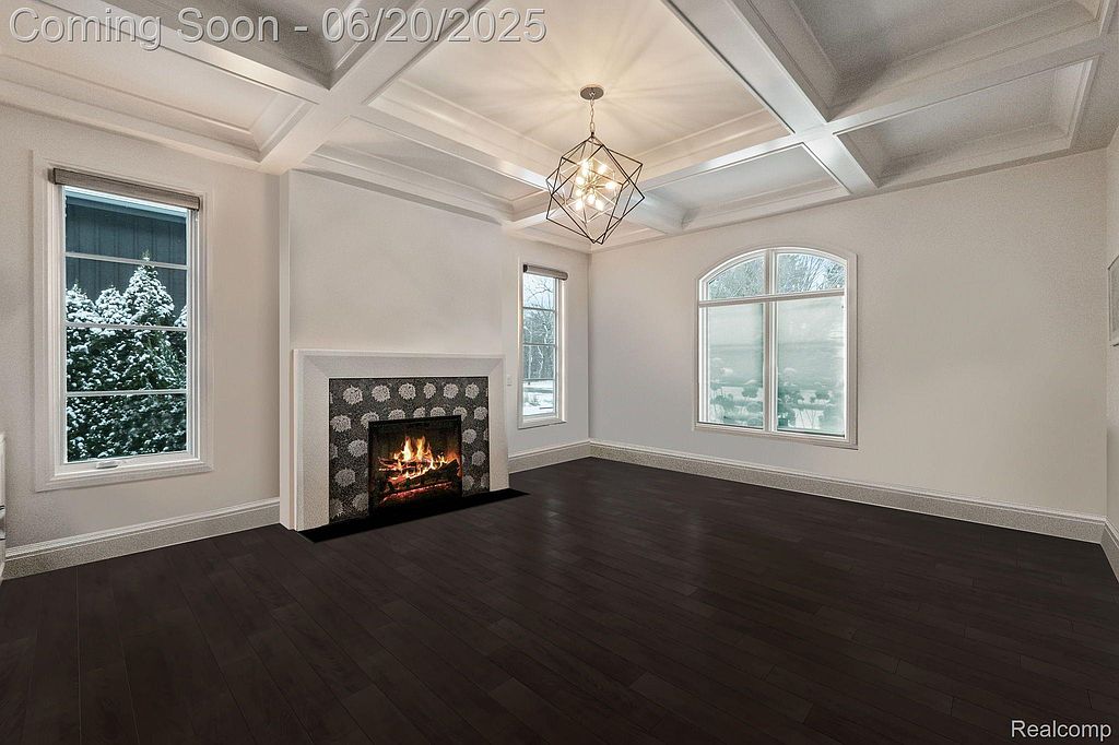 This is an interior shot of a living room featuring dark hardwood floors, a coffered ceiling with a modern chandelier, and a fireplace with a decorative surround. Large windows provide natural light, creating a bright and inviting space. The room appears spacious and well-appointed, suitable for entertaining or relaxing.