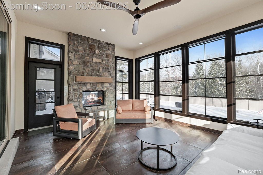 This sunroom features a stone fireplace, large windows offering ample natural light and views of the snowy outdoors, and comfortable seating. The dark-framed windows and tile flooring add a touch of modern elegance, creating a cozy and inviting atmosphere. A ceiling fan provides air circulation.