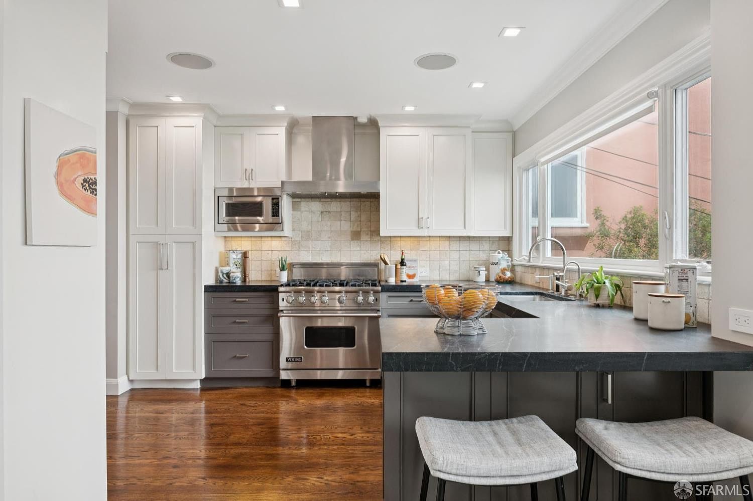 This is a well-lit kitchen featuring a stainless steel Viking range and hood, white upper cabinets, and gray lower cabinets. The countertops are dark, possibly granite or a similar material, and there's an island with bar seating. A window above the sink provides natural light, and the hardwood floors add warmth to the space. The perspective is from a medium distance, showcasing the layout and key features of the kitchen.