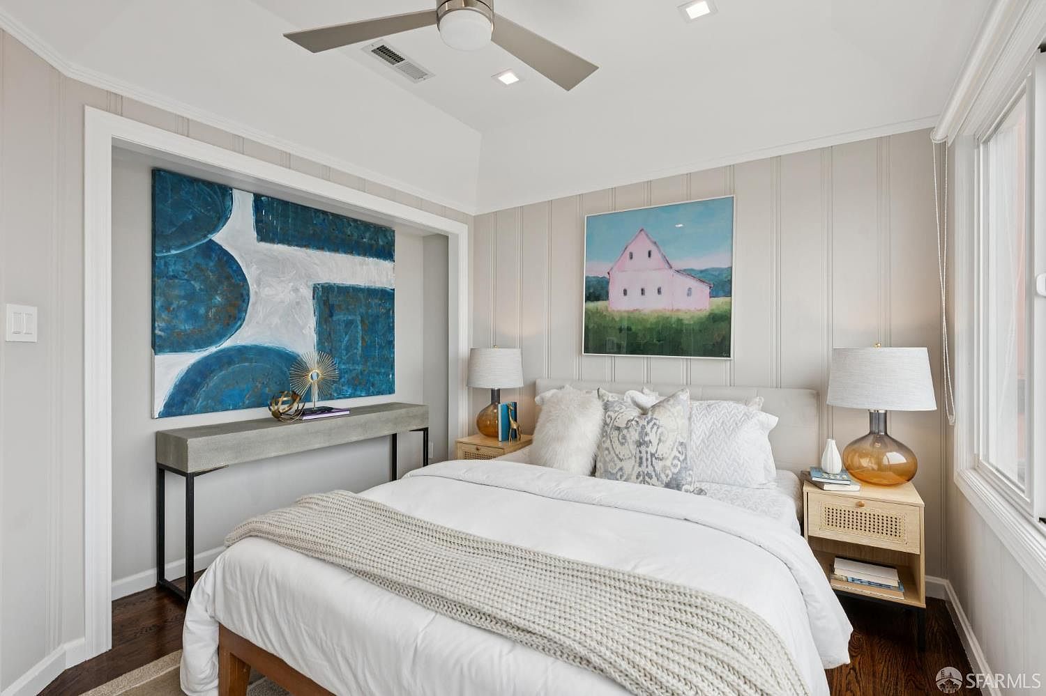 This is a well-lit primary bedroom featuring a bed with white linens and a gray throw blanket. The room is decorated with artwork above the bed and a console table, as well as on the adjacent wall. Nightstands with lamps flank the bed, and the walls are paneled in a neutral tone, creating a serene and inviting atmosphere.