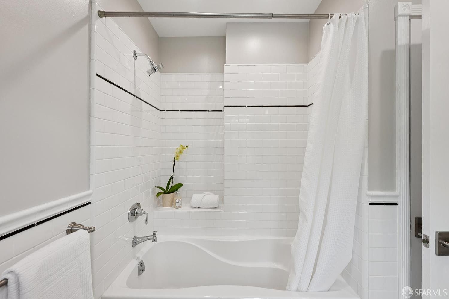 This is a well-lit bathroom featuring a white bathtub with white subway tile surround and a shower curtain. A black accent stripe runs horizontally through the tiling, adding a touch of contrast. A small shelf holds a potted plant and a neatly folded towel, contributing to a clean and inviting atmosphere.