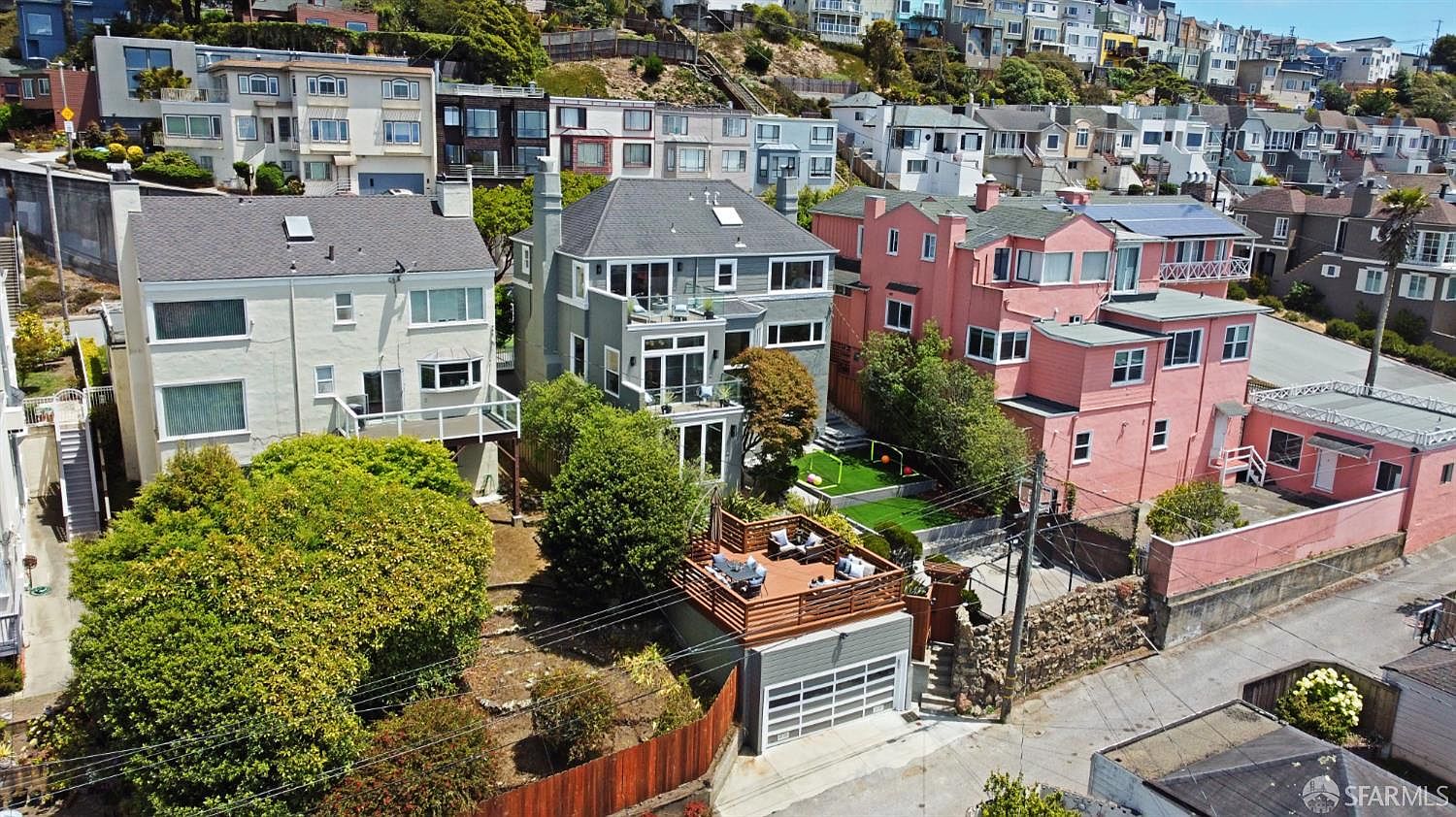 This aerial shot showcases a residential neighborhood with a focus on a multi-level home featuring a gray exterior, multiple balconies, and a well-maintained backyard with a small lawn and a deck above the garage. The surrounding houses are a mix of styles and colors, nestled on a hillside, creating a dense yet appealing residential landscape.