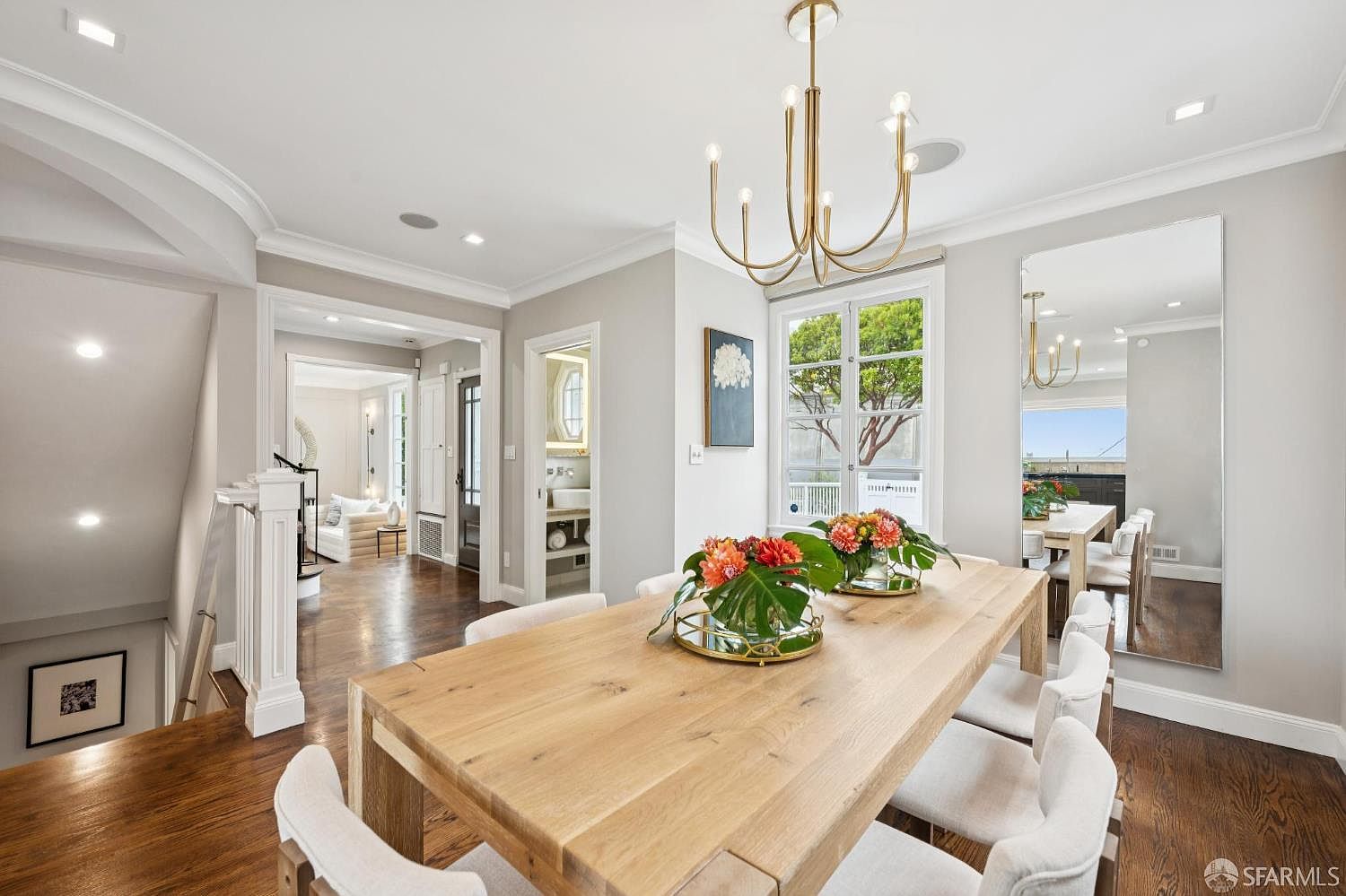 This is an interior shot of a dining room featuring a large wooden table with white upholstered chairs. A modern chandelier hangs above the table, and a large window provides natural light. The room has a neutral color palette and hardwood floors, creating a warm and inviting atmosphere.