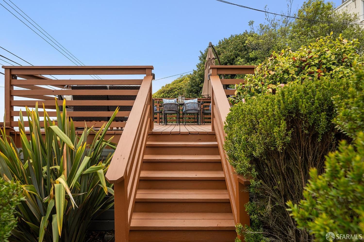 This image showcases a charming outdoor deck accessible via wooden stairs. The deck features patio furniture, suggesting a comfortable space for relaxation and entertainment. Lush greenery surrounds the deck, enhancing its appeal and creating a sense of privacy.