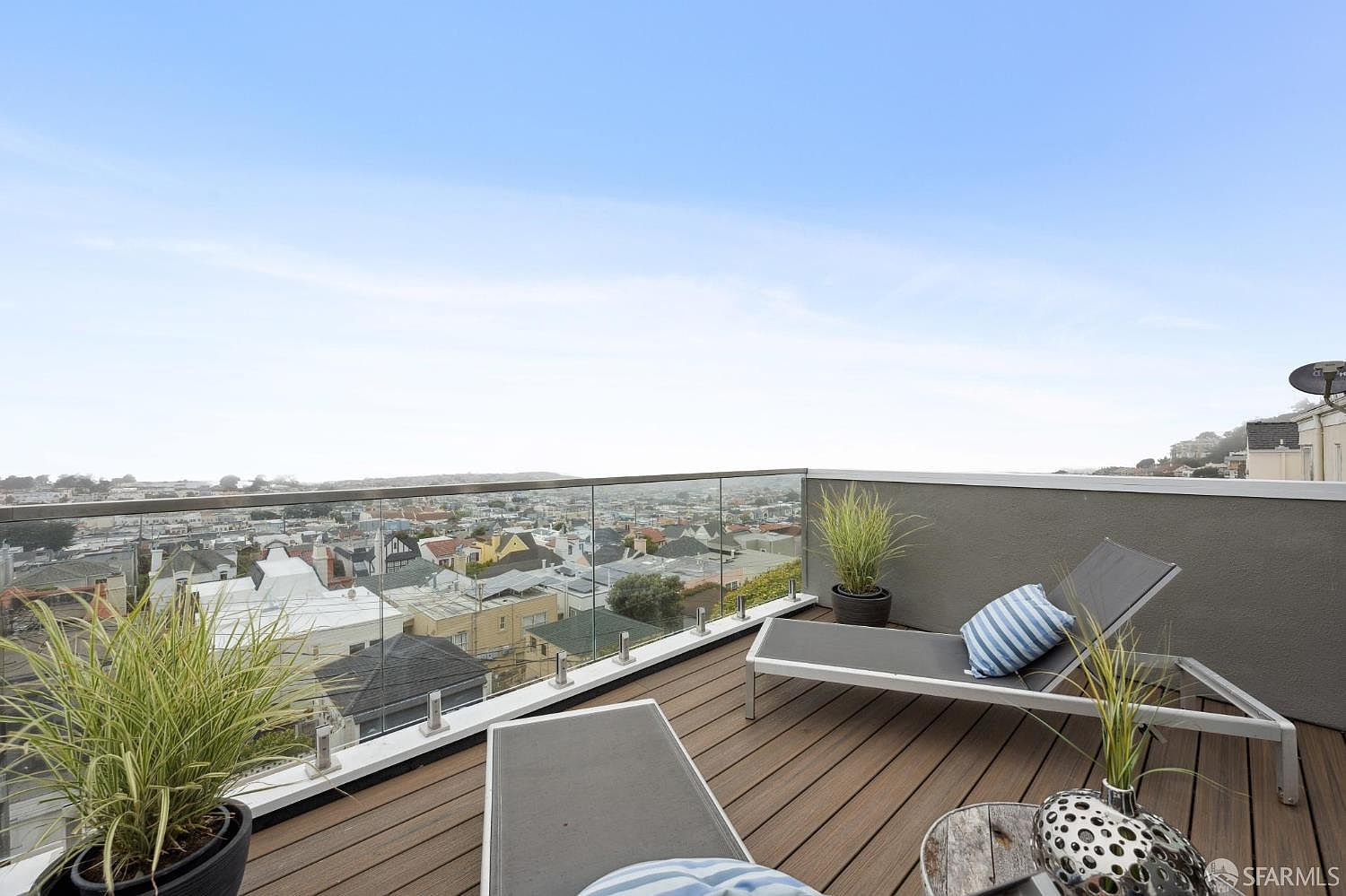 This image showcases a stylish rooftop deck with a stunning panoramic city view. The deck features modern lounge chairs, potted plants, and a sleek glass railing that maximizes the vista. The wooden decking adds warmth and texture, creating an inviting outdoor living space perfect for relaxation and entertaining.
