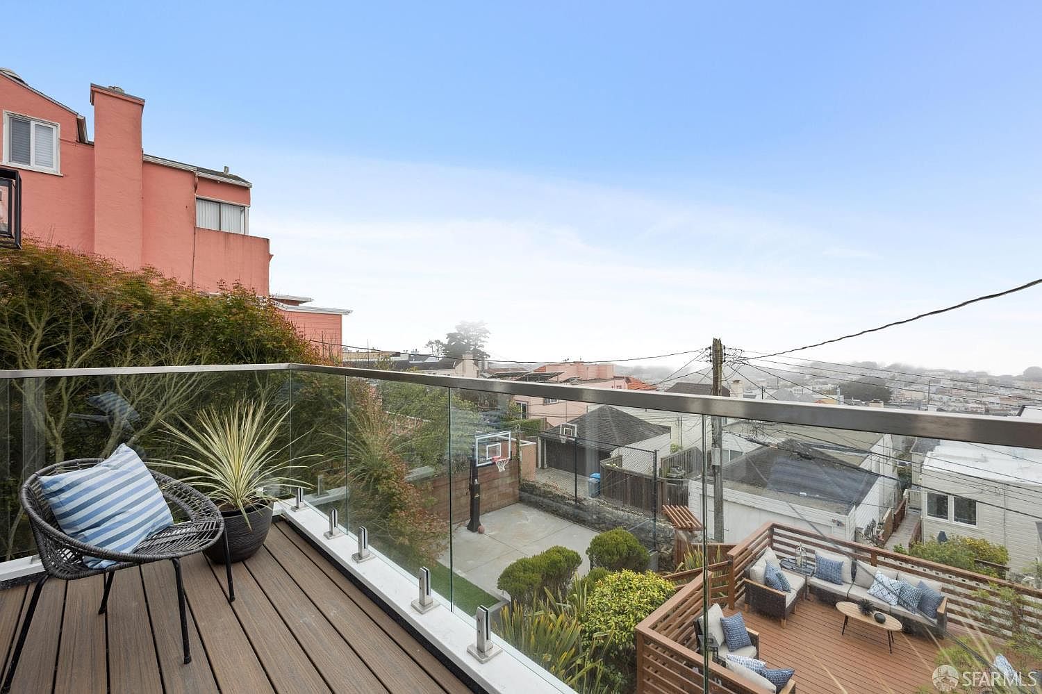 This image showcases a stylish outdoor living space, featuring a modern deck with glass railings that offer unobstructed views of the surrounding neighborhood. The deck is furnished with comfortable seating arrangements, including a lounge area and a cozy chair with a striped pillow. The space is well-maintained with lush greenery, creating a relaxing and inviting atmosphere.