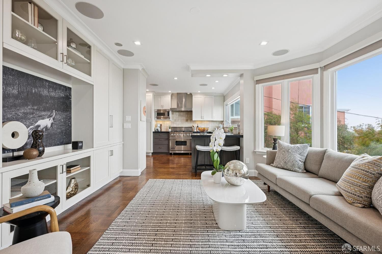 This is an interior shot of a living room that flows into a kitchen. The living room features a neutral-toned sofa, a modern white coffee table, and a patterned rug. Built-in shelving and cabinetry provide ample storage, while large windows offer natural light and a view of the outside. The overall impression is a bright, open, and inviting living space.