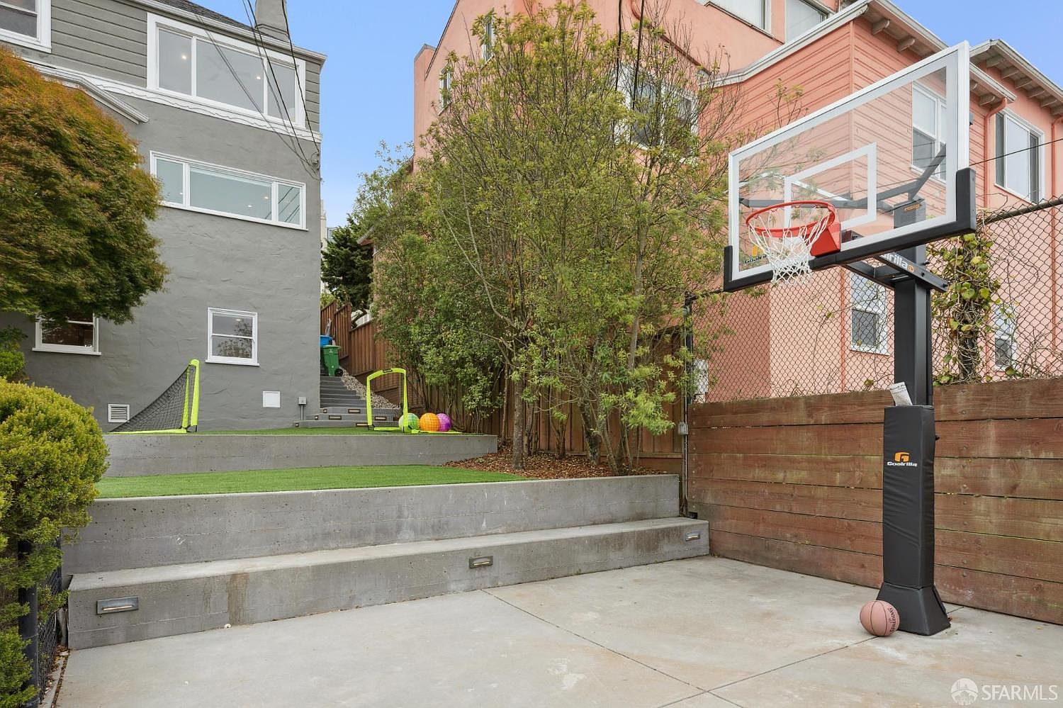 This image showcases a well-maintained backyard featuring a basketball court and a grassy area with soccer goals. The yard is tiered with concrete retaining walls and includes mature trees and landscaping. The presence of recreational elements suggests a family-friendly environment, perfect for outdoor activities and relaxation.